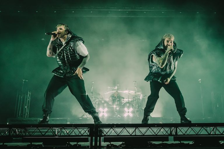 Electric Callboy frontmen performing live on stage in matching black puffy vests under green atmospheric lighting.